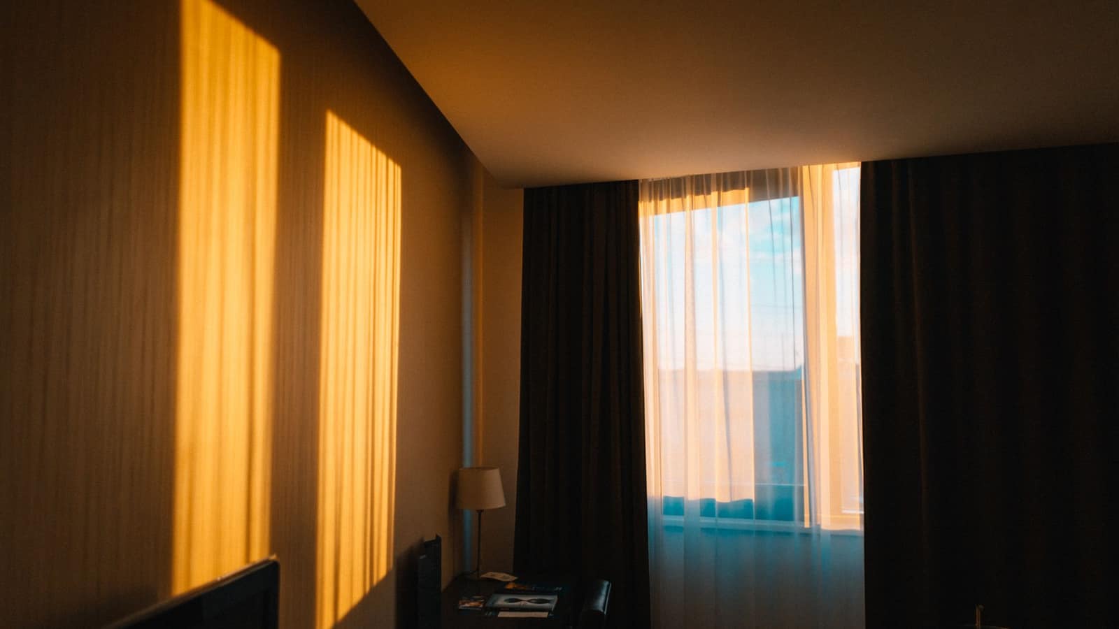 Warm lamp light filling a bedroom beside a curtained window at dawn